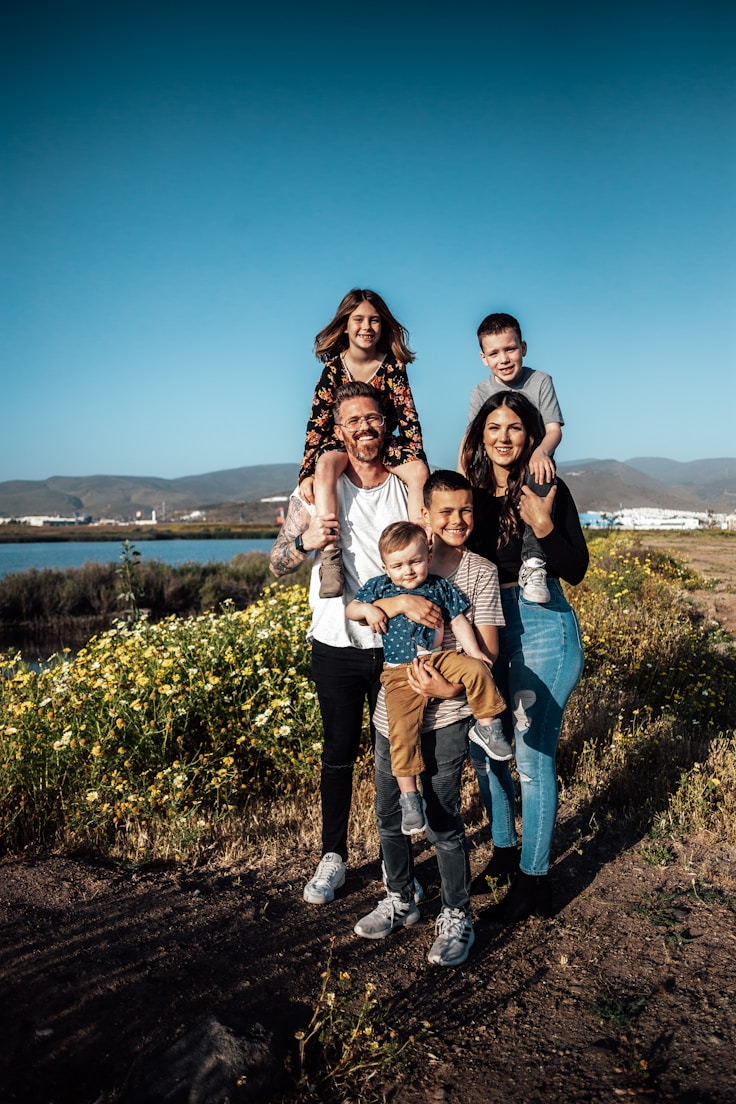 Smiling diverse family of six with kids on shoulders amid wildflowers and lagoon, illustrating family law and child welfare services.